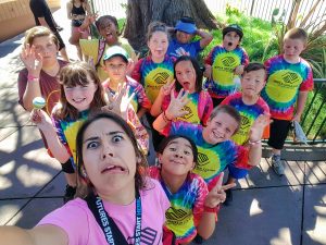 A group of smiling and playful kids in colorful tie-dye shirts pose for a selfie with a young woman, all making funny faces and gestures outside by a large tree on a sunny day.