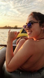 A woman wearing sunglasses smiles while sitting in an inflatable boat on the water at sunset, with sunlight shining on her face and a scenic shoreline in the background.