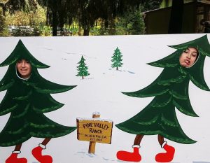 Two people pose with their faces through cutouts in a painted board of walking Christmas trees at Pine Valley Ranch in Auburn, CA. The board also features cartoon trees, a wooden sign, and trees in the background.