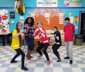 Five young people pose playfully in a colorful youth center. Three are dressed as Power Rangers, one wears a plaid shirt and hat, and one in a red polo shirt stands smiling on the right. Signs and posters decorate the walls behind them.