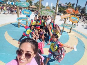 A group of kids in colorful tie-dye shirts poses for a cheerful selfie at a water park, with signs and crowds in the background on a sunny day.