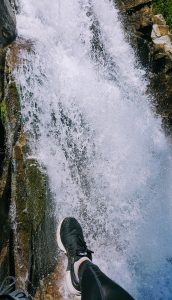 A person’s leg in black athletic clothing and a black sneaker extends over the edge of a rocky waterfall, with rushing white water cascading down below.