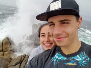 A young couple smiles for a selfie in front of ocean waves crashing against rocks; the man wears a dark hat and the woman is behind him, both looking happy.