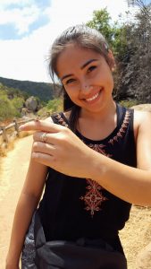 A smiling young woman outdoors on a sunny day holds out her hand to show a small insect perched on her finger. She wears a black sleeveless top with an embroidered design and stands on a dirt path surrounded by trees and rocks.