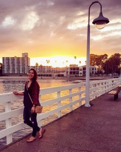 A woman stands smiling on a pier at sunset, with a lamp post nearby, buildings, palm trees, and colorful clouds in the background. The scene overlooks calm water and has a relaxed, warm atmosphere.