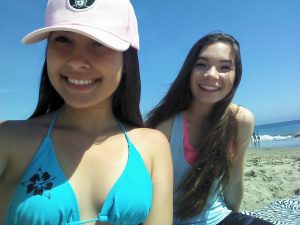 Two young women smiling at the beach. One wears a pink cap and blue bikini top with a flower design, while the other has long hair and wears a light blue tank top. The sky is clear and the ocean is in the background.