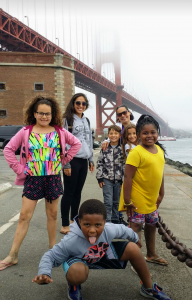 A group of smiling adults and children pose playfully near the Golden Gate Bridge on a foggy day, with some kids making silly faces and gestures in the foreground.
