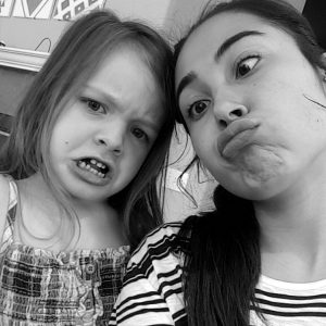 A young girl and a woman take a black-and-white selfie, both making funny faces; the girl frowns dramatically, while the woman pouts her lips and widens her eyes.