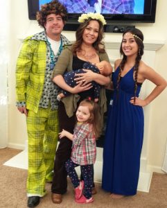 A family poses indoors in costume: a man in a bright plaid suit, a woman with a flower crown holding a baby, a little girl in plaid, and a young woman in a blue dress with a headband and long braids.