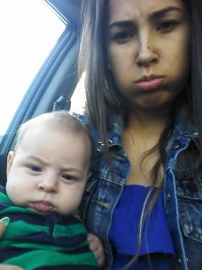 A woman with long brown hair and a denim jacket puffs out her cheeks, making a funny face, while holding a baby in a green and navy striped outfit who is also making a serious expression. They are sitting in a car.