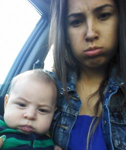 A woman and a baby sit together in a car, both making playful pouty faces. The woman wears a denim jacket and has long brown hair, while the baby wears a green and black striped outfit.
