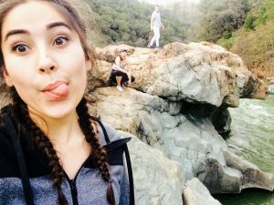 Young woman with dark braided hair makes a silly face in the foreground; behind her, two people stand and crouch on large rocks near a river, with trees and greenery in the background.