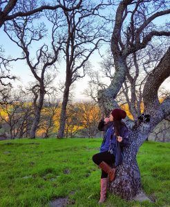 A woman in a maroon beanie and boots leans against a tree on a grassy hill, surrounded by bare-branched trees, with golden light illuminating the background.