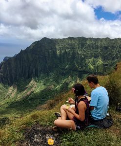 Two people sit on a grassy cliff edge having a picnic, overlooking a lush green mountain range under a partly cloudy sky. The scene is peaceful and scenic, with food containers and drinks beside them.