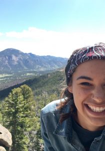 A smiling person with a nose ring and patterned headband poses in front of a scenic mountain landscape with green trees and clear sky.