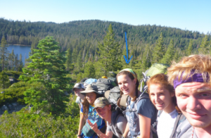 Six young people with backpacks are hiking in a forested area near a lake, posing and smiling at the camera. One person in the middle is marked with a blue arrow above their head.