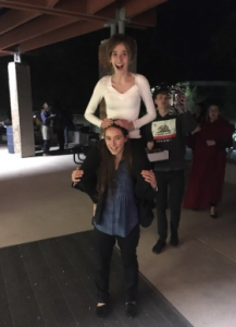 Two young women are smiling; one is sitting on the other’s shoulders. They are outside at night under a covered area, with other people standing and walking in the background.