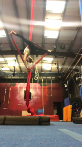 A woman performs an aerial silk pose, hanging upside down with her legs extended in a split while gripping red fabric in a gym with ropes and mats visible in the background.