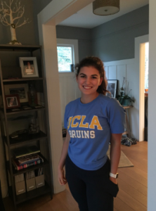 A smiling woman stands indoors wearing a blue UCLA Bruins t-shirt and dark pants, with a bookshelf and framed photos in the background.