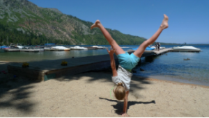 A child in a white shirt and blue shorts does a cartwheel on a sandy beach near a lake, with docks and boats in the background and forested hills under a clear blue sky.