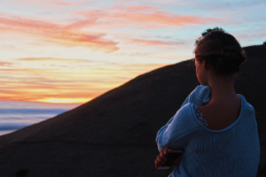 A woman with her hair tied back, wearing a light blue sweater, stands with arms crossed, holding a phone, and looks at a colorful sunset sky over a rocky landscape.