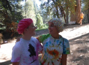Two people wearing colorful shower caps and tie-dye shirts stand outdoors in a wooded area, smiling at each other with arms around each others shoulders. Sunlight filters through the trees in the background.