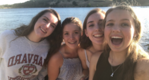 Four young women smile and pose for a selfie by a lakeside on a sunny day. One wears a gray Harvard sweatshirt, while the others wear light summer tops. Trees and hills are visible in the background.