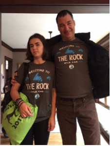 A man and a young girl stand indoors, both wearing matching grey Welcome to The Rock t-shirts. The girl holds a green drawstring bag, and the man has his arm around her shoulders while smiling.