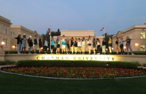 A group of people stand in a row with arms outstretched in front of an illuminated Chapman University sign, with university buildings and flowerbeds in the background at dusk.