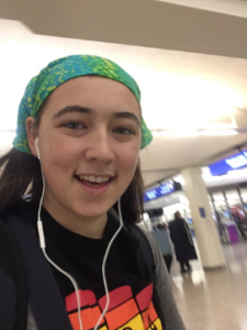 A young person smiling at the camera, wearing a green and blue bandana, earphones, and a black shirt with a colorful design, standing in what appears to be an airport terminal.