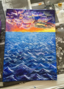 A painting shows a vibrant sunset sky with purple, orange, and yellow clouds above a choppy blue ocean, with white highlights on the waves. The canvas rests on top of newspaper.