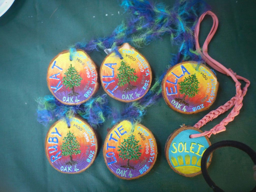 Five colorful wooden name tags with painted trees and names (Ruby, Kat, Ollie, Ella, Solei) on yarn or ribbon lanyards are arranged on a green surface. The tags also say “Camp Augusta 2018” and OAK.