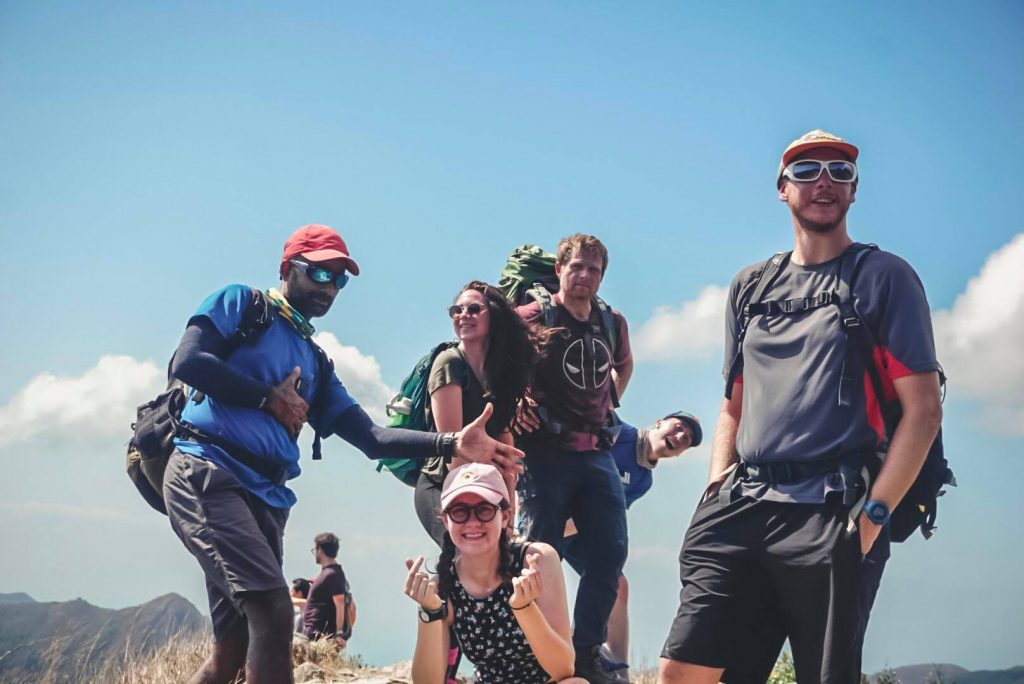 A group of six hikers with backpacks pose and smile on a mountain summit under a clear blue sky. They wear hats and sunglasses, with mountains and clouds visible in the background.