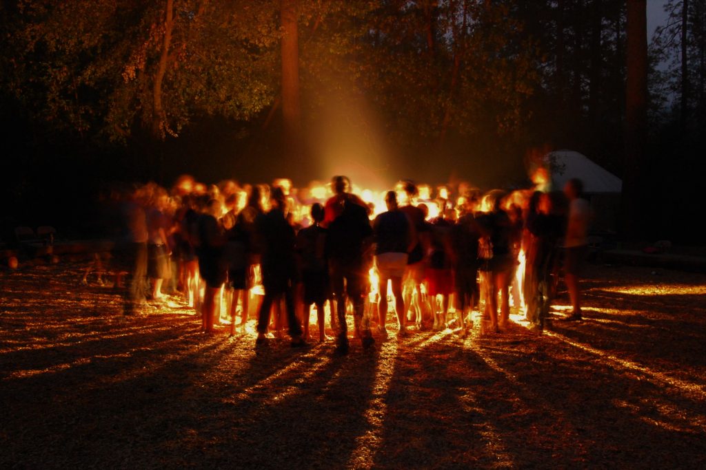 A large group of people stand in a circle around a glowing campfire at night, casting long shadows on the ground in a wooded outdoor setting. Trees and a white tent are visible in the background.
