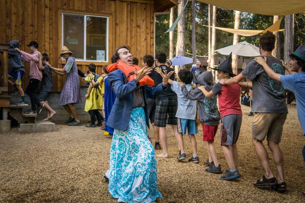 A group of children and teens form a conga line outside near a wooden building in a forested area. One person in front, wearing a blue patterned skirt and hoodie, leads with an animated expression. Some wear costumes or hats.