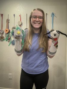A smiling woman with long blonde hair and glasses holds two sock puppets, one resembling a green creature and the other a black-and-white dog, standing in front of a wall decorated with colorful medals and ukuleles.