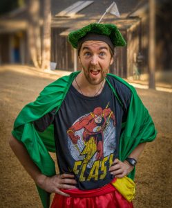 A man in a green cape, grass-like hat, and red shorts stands with hands on hips, wearing a Flash superhero t-shirt and smiling with a surprised expression outdoors.