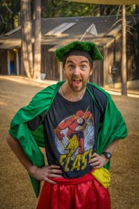 A man stands outdoors wearing a green cape and grass-like hat, with a Flash superhero t-shirt and red shorts. He is posing playfully with hands on hips and a surprised expression, with cabins and trees in the background.