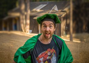 A man wearing a green cape and a hat made of artificial grass with a small flag on top stands outdoors, looking excited. He is also wearing a Flash superhero T-shirt. A wooden building is blurred in the background.