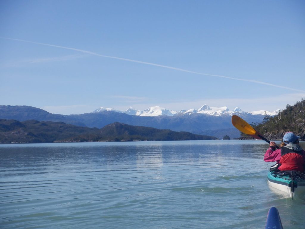Two people paddle a kayak on a calm lake with clear blue water, surrounded by green hills and distant snow-capped mountains under a blue sky.