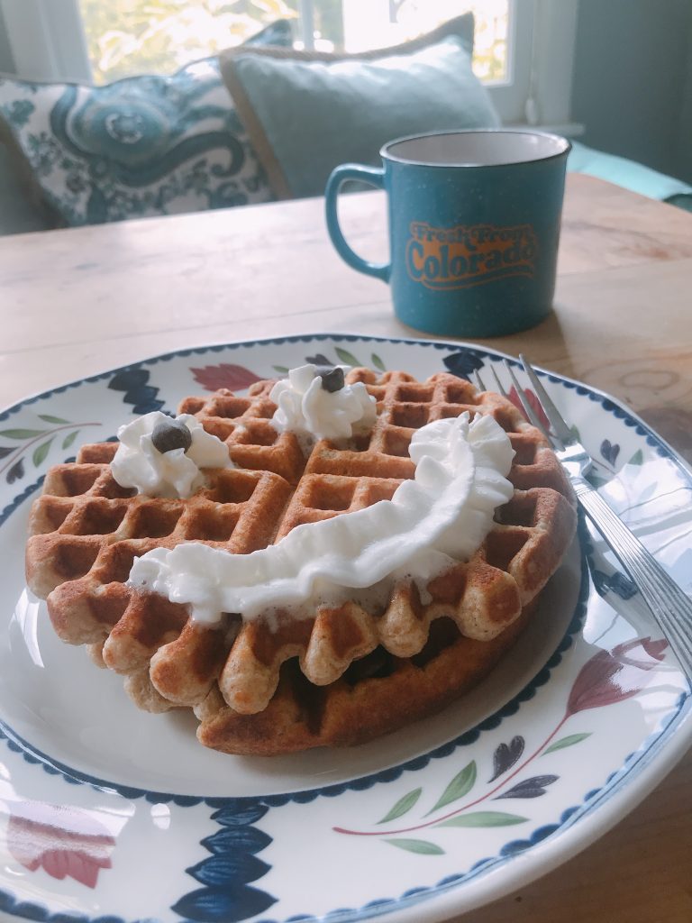 A waffle on a decorative plate is topped with whipped cream and chocolate chips arranged as a smiling face. A fork and knife are beside the plate. In the background, there’s a blue Colorado mug and pillows near a bright window.