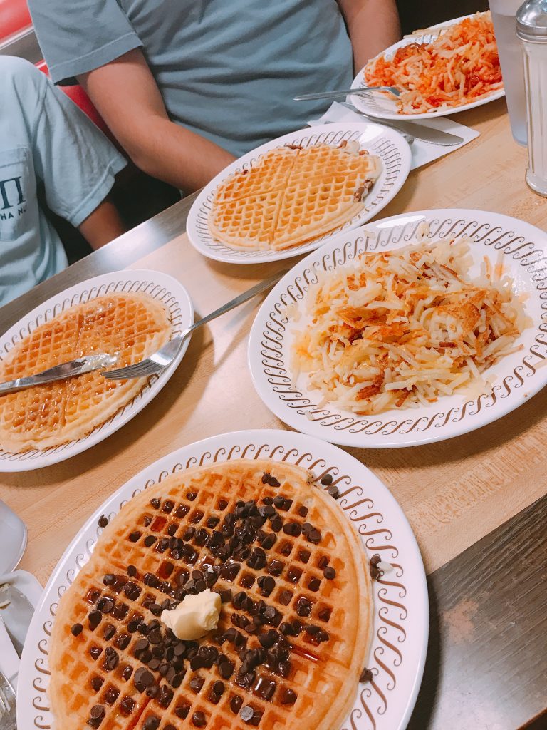 Four plates of waffles, one topped with chocolate chips and butter, and a plate of hash browns are set on a diner table. Two people are seated, partially visible, ready to eat.