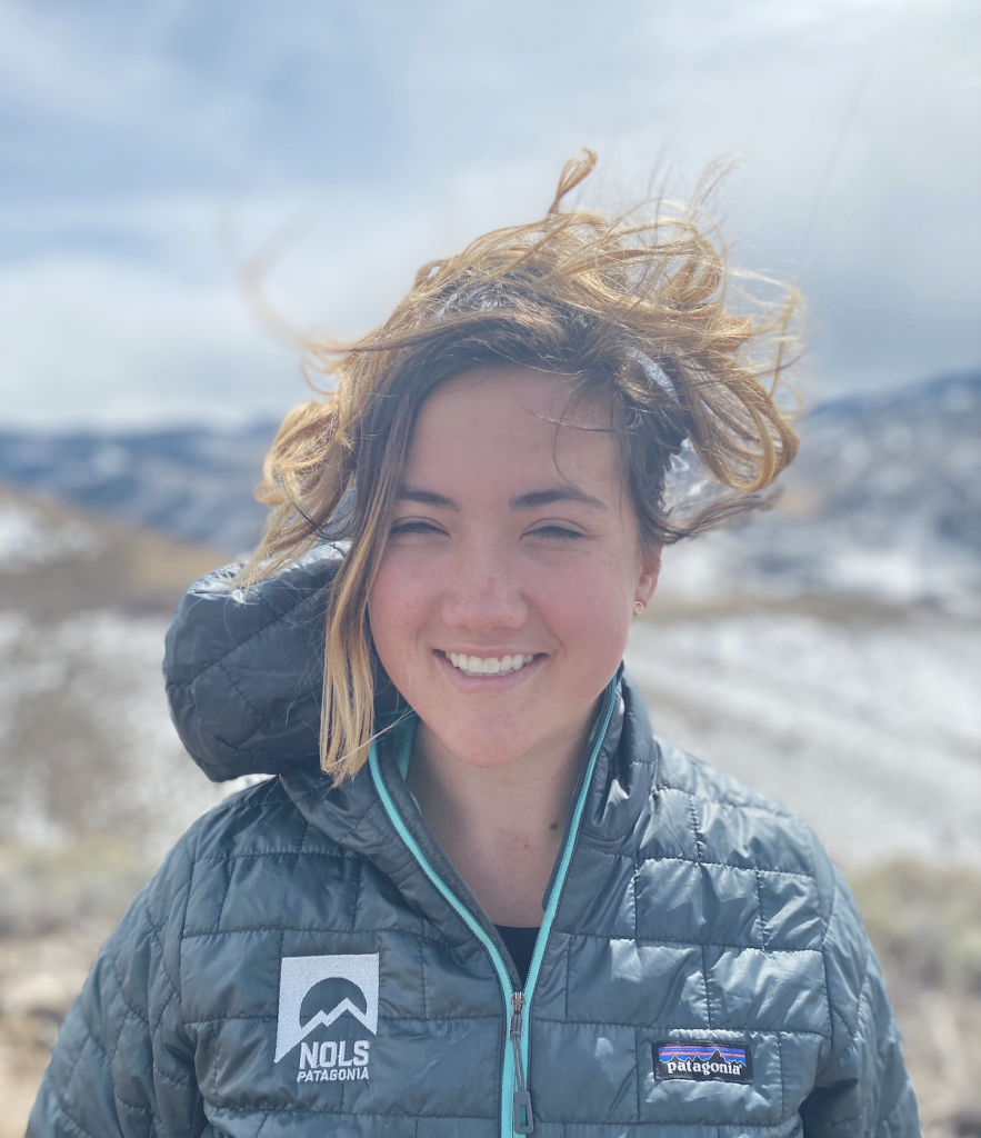 A woman smiles outdoors on a windy day, her hair tousled by the wind. She wears a gray NOLS Patagonia jacket, with snowy mountains and a cloudy sky in the background.