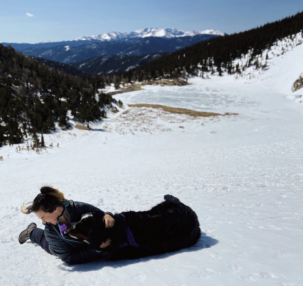 A woman sits and cuddles a large black dog on a snowy hillside, with a frozen lake, forested slopes, and distant mountains under a blue sky in the background.