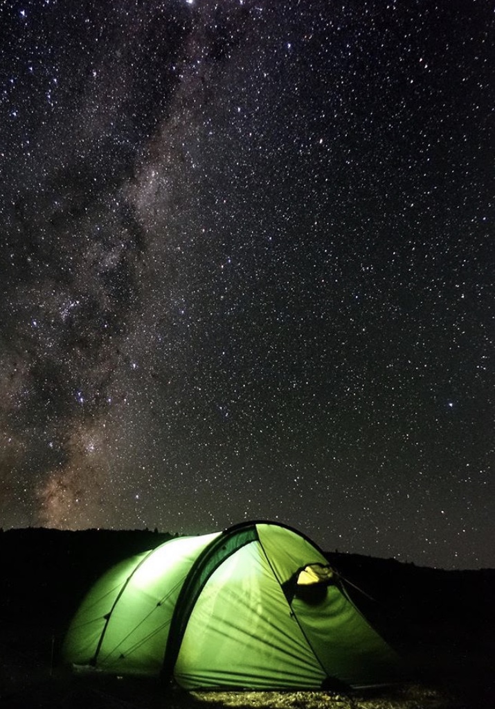 A glowing green tent is pitched under a dark, star-filled night sky with the Milky Way visible. The tent is on a flat landscape, and the stars create a serene, awe-inspiring atmosphere.