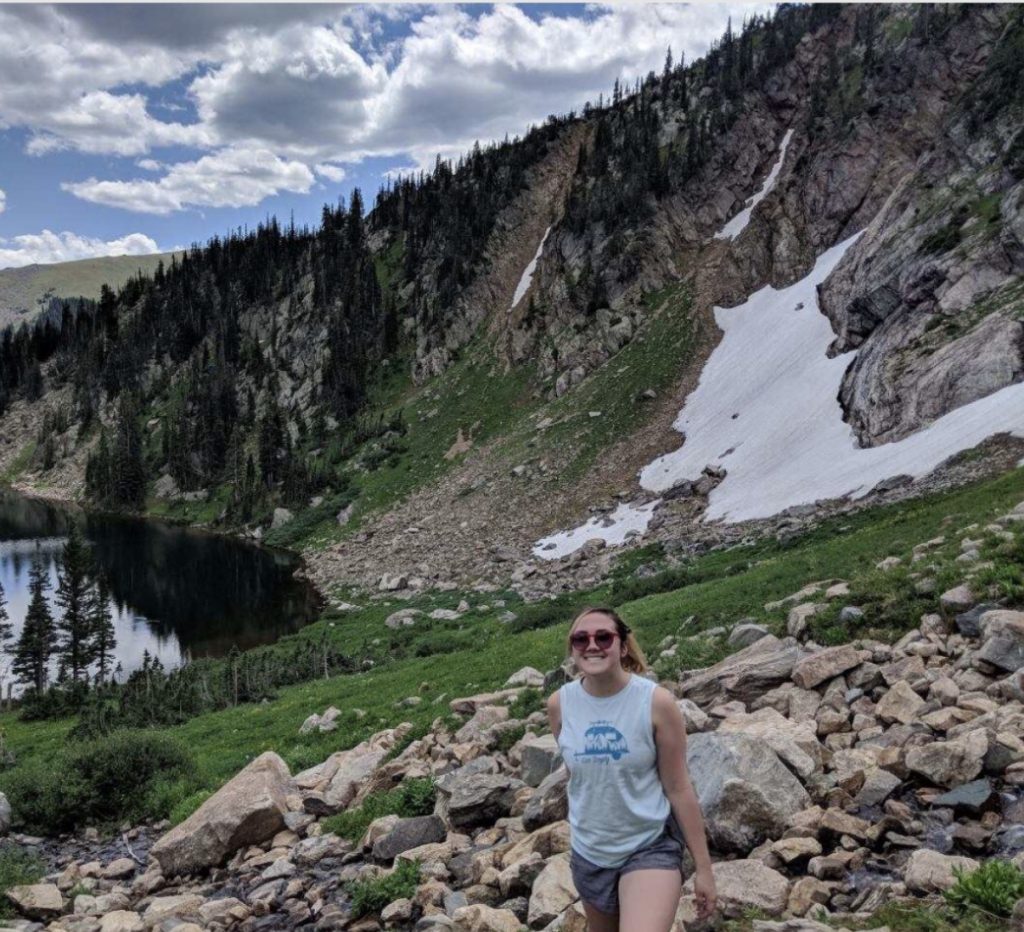 A woman in sunglasses and a sleeveless shirt smiles while standing on rocky terrain by a mountain lake, with a forested slope and patches of snow in the background under a partly cloudy sky.