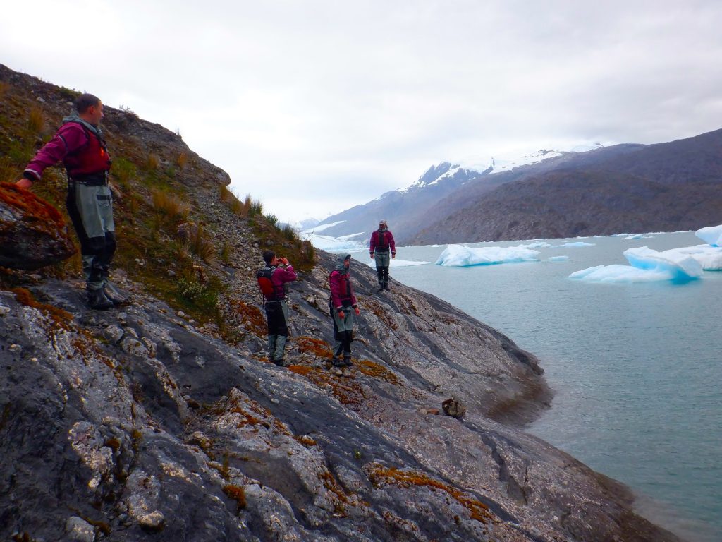 Four people in outdoor gear stand on a rocky slope beside a lake with floating icebergs. Snow-capped mountains are visible in the background under a cloudy sky.