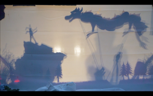 A shadow puppet scene shows a figure standing on a rock facing a large dragon, with visible rods controlling the dragon. Trees and other shadowy figures are in the background against a light-colored backdrop.