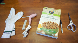 A pair of white tube socks, a hot glue gun with glue sticks, a Sharpie marker, a box of organic Crunchy Cinnamon Squares cereal, and a pair of scissors are arranged on a wooden table.