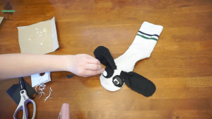 A hand attaches a black felt circle to a sock craft shaped like a dogs face, with black ears and paper eyes, on a wooden table with scissors, cardboard, and craft supplies nearby.
